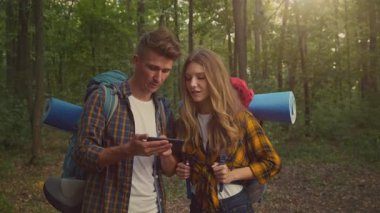 Bir çift, canlı bir ormanda yürüyüş yapmaya ara verdiklerinde akıllı telefonlarıyla meşgul olurlar. Sırt çantaları arkalarında duruyor ve güneş ağaçların arasından süzülüyor..