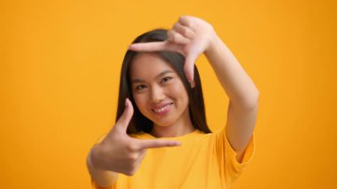 Genç bir Asyalı kadın, neşeli bir şekilde elleriyle yüzünü çerçeveliyor, canlı sarı bir arkaplana karşı yaratıcılık sergiliyor. İfadesi özgüven ve kendini ifade etme hevesi yayıyor..