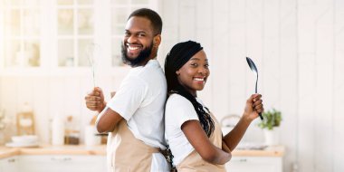 Birlikte yemek pişirirken önlük takan neşeli siyah eşlerin portresi, Afrika kökenli Amerikalı erkek ve kadının elinde mutfak gereçleri tutması ve kameraya gülümsemesi.