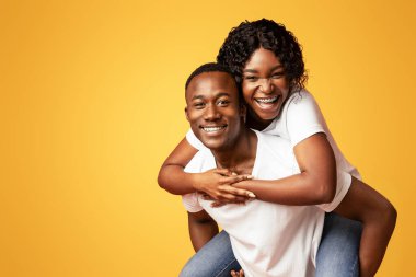 Mutlu siyahi aşıklar genç adam ve kadın turuncu stüdyo arka planında poz veriyorlar, kopyalama alanı. Neşeli Afrikalı Amerikalı kadın erkeğini sırtında taşıyor, reklam için boş bir alanda duruyor.