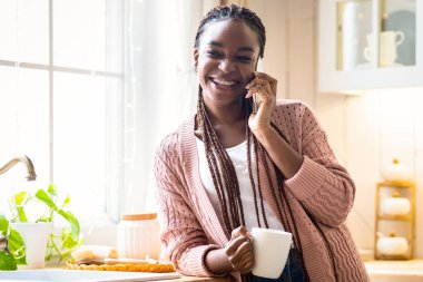 Yurt içi eğlence. Neşeli Afrikalı Amerikalı kadın kahve içiyor ve cep telefonuyla konuşuyor, siyah kadın cep telefonuyla konuşuyor ve mutfak içinde sıcak içkinin tadını çıkarıyor, bedava uzay...