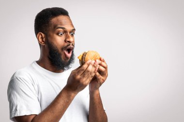 Heyecanlı Siyah Adam Sağlıksız Burger Poz Veriyor Beyaz Stüdyo Arkaplanında Duruyor. Abur cubur yiyici ağzı açık, lezzetli, sağlıksız hamburgerciye bakıyor. Beslenme ve aşırı yeme alışkanlığı