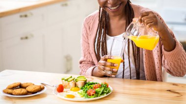Neşeli Afro-Amerikalı kadın evdeki mutfakta sağlıklı ve lezzetli bir kahvaltı yiyor. Pozitif siyah milenyum kadını masada oturuyor, lezzetli yemeklerin tadını çıkarıyor ve portakal suyu içiyor, fotokopi çekiyor.