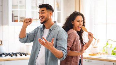 Ev Konseri. Neşeli Orta Doğulu eşler mutfakta eğleniyor, şarkı söylüyor ve dans ediyor. Neşeli Arap Çifti Spatulaları Mikrofon Olarak, Modern Müslüman Aile Evde Yemek Pişirirken Kandırıyor