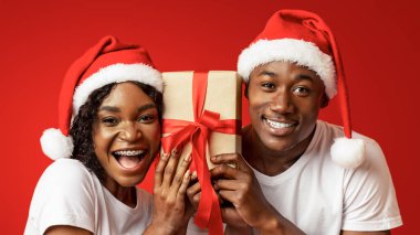 Noel, yeni yıl kutlaması. Noel Baba şapkalı duygusal siyah çift 2021 'i birlikte kutluyorlar, hediye kutusunu bir arada tutuyorlar ve kameraya gülümsüyorlar, turuncu stüdyo arka planı, yakın çekim.