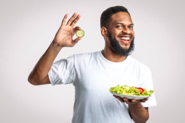 Sebzeli salata tutan neşeli siyah adam sağlıklı yiyecekler seçiyor, beyaz stüdyonun arkasında duruyor, kameraya gülümsüyor. Zayıflatıcı Beslenme ve Erkek Kilosu Kaybı