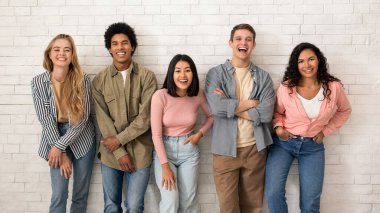 Öğrenci hayatı, arkadaşlık ve modern öğrenim değişimleri hakkında blog yaz. Neşeli genç çok ırklı öğrenci takımı gündelik olarak eğlenin ve kameraya bakın, beyaz tuğlalı arka planda, stüdyo çekimleri