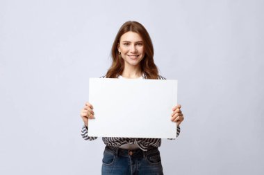 Reklam için fotokopi alanı olan boş pankart tutan güzel genç bir kadın. Gülümseyen milenyum hanımefendisi, beyaz boş ilan panosu ve yazı için yer gösteriyor, gri stüdyo arka planının başında duruyor.