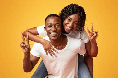 Mutlu siyahi aşıklar genç adam ve kadın turuncu stüdyo arka planında poz veriyorlar, kopyalama alanı. Neşeli Afrikalı Amerikalı kadın erkeğini sırtında taşıyor, barış jestleri yapıyor, yakın plan