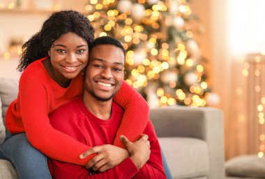 Covid-19 karantinasında romantizm ve yeni yıl kutlaması. Mutlu, genç, Afro-Amerikan karı-koca kucaklaşıp, Noel ağacının parıldayan çelenkleriyle evlerinde kameralara bakıyorlar.