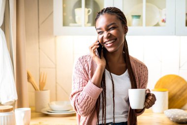 Neşeli siyah kadın cep telefonuyla konuşuyor ve mutfakta kahve içiyor. Pozitif siyah kadın elinde bardakla sıcak içecek tutuyor ve arkadaşlarıyla hoş sohbetler ediyor.