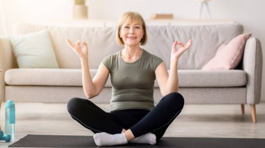 Lotus poz veren dengeli yaşlı kadın elleriyle Gyan Mudra yapıyor, evdeki spor paspası üzerinde meditasyon yapıyor. Huzurlu olgun bayan yoga ve farkındalık yapıyor, kopyalama alanı