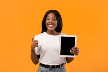 Yeni dijital tablet gösteren güzel siyahi bir kadın. Boş ekran ve baş parmağı yukarıda. Turuncu stüdyo arka planı, kopyalama alanı. Mutlu Afro-Amerikalı kadın, boş ekranlı, maketli.