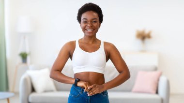 Başarılı Kilo Verme. Sıska kot pantolon giyen, evinde dikilip kameraya gülümseyen, zayıf gösteren mutlu Afrikalı kadın. Kilo Verme Motivasyonu Konsepti