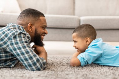 Babalar Günü. Afrikalı babayı sevmenin ve küçük oğlunun evde yerde yatarken birbirlerine bakmalarının yan görüntüsü. Genç baba ve oğlu kapalı alanda kaynaşıyor. Baba ve Çocuk, Aile Kavramı