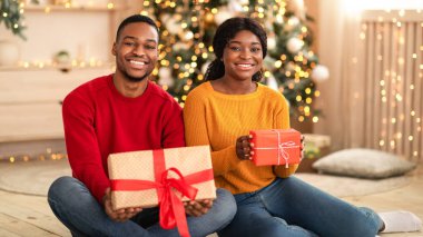 Gifts for family and New Years home decor. Millennial happy african american couple sit on floor and hold boxes with present in living room interior with Xmas tree and glowing garlands, free space