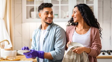 Neşeli Orta Doğulu çift ev işlerini paylaşıyor, mutfakta birlikte bulaşık yıkıyorlar, mutlu Arap eşler evde temizlik yapmaktan zevk alıyorlar, birbirlerine bakıp gülümsüyorlar.