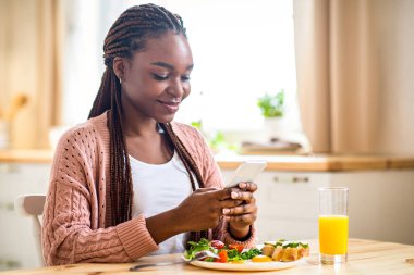 Mutlu Siyah Kadın Kahvaltı Yapıyor ve Akıllı Telefonu Mutfakta Kullanıyor, Neşeli Afrikalı Afrikalı Amerikalı Kadın Sosyal ağlara ya da Mesajlara Bakıyor Sabah Yemeğinin, Boş Alan 'ın keyfini çıkarırken