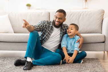 Afrikalı baba ve oğul yerde otururken, baba çocuğunu evde gösteren bir şeyi parmağıyla işaret ediyor. Aile Mutluluğu, Mutlu Babalık ve Çocukluk Konsepti