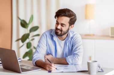 Belgeler ve bilgisayarla çalışan başarılı bir Doğu girişimcisi, bilgisayar ekranına bakan gülümseyen Arap iş adamı, mali raporları kontrol eden, not alan, boş alan...