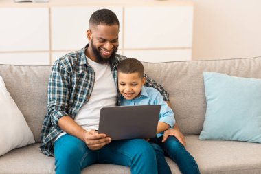 Afrikalı baba ve oğul dizüstü bilgisayarda çizgi film izliyorlar evde koltukta birlikte oturuyorlar. Babamla hafta sonu. Aile Yaşam Tarzı ve Gadget Kavramı