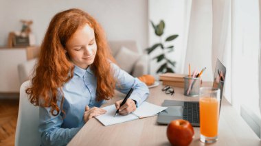 Uzaklık Eğitim Konsepti. Masasında dizüstü bilgisayarıyla oturan ve defterine ya da günlüğüne yazan gülümseyen genç kız. Öğrenci kız çevrimiçi ders izliyor, not alıyor, ödev yapıyor.