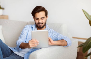 Yakışıklı Doğulu adam evde dinlenirken dijital tabletle internette geziniyor. Oturma odasında oturan gülümseyen Arap genç adam, sekme bilgisayarını kullanıyor, internetten alışveriş yapıyor ya da internette sörf yapıyor.