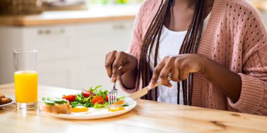 Kahvaltı vakti geldi. Afro-Afrikalı Bayan, mutfakta sağlıklı besleniyor, çatal ve bıçak kullanırken gülümsüyor, lezzetli yemeklerin tadını çıkarıyor, taze salata, yumurta ve sandviç yiyor,