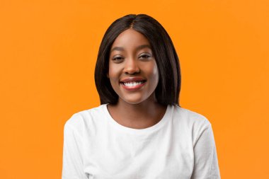 Turuncu stüdyo arka planında poz veren beyaz tişörtlü, sevimli, siyah bir bayanın yakın plan portresi, fotokopi alanı. Güzel Afro-Amerikalı kadın. Parlak sarı saçlı. Kameraya gülümseyen, flört eden.