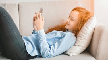 Dijital Yerliler. Cep telefonunu kullanarak gülümseyen, internetten video izleyen, dinlenen ve evdeki oturma odasındaki kanepede uzanan genç kızın portresi. Çocuk tek başına eğleniyor.