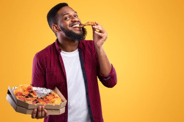 Pizzacı. Komik siyah adam abur cubur yiyor, pizza dilimleri yiyor, sarı stüdyo arka planında pizza kutusuyla poz veriyor, kameraya gülümsüyor. Sağlıksız Beslenme Alışkanlıkları, Aldatma Yemekleri