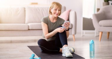 Son sınıf öğrencisi kadın, antrenmanından sonra yoga minderinde akıllı telefonuyla rahatlıyor ve internetteki yeni spor videolarına bakıyor. Yoga antremanından sonra mola veren olgun beyaz kadın. Etkin yaşam tarzı kavramı