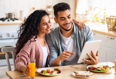 Neşeli Orta Doğulu eşler kahvaltı yaparken dijital tablet kullanıyorlar. Modern Gadget ile masaya oturan, internetten alışveriş yapan ya da interneti tarayan mutlu genç bir Arap çift.