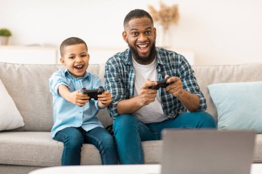 Siyah küçük çocuk ve babası evde video oyunu oynuyorlar. Birlikte eğleniyorlar. Genç baba oğluyla oyunlar oynuyor. Çocuklar ve aileler için video oyunları, hafta sonu eğlence konsepti. Seçici Odaklanma