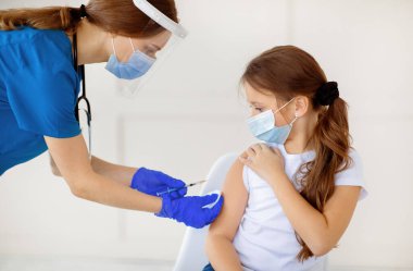 Covid-19 aşı kampanyası. Koruyucu maskeli küçük bir kız hastanedeki doktordan koronavirüs aşısı alıyor. Antiviral bağışıklık sistemi, bulaşıcı hastalıkları önleme konsepti