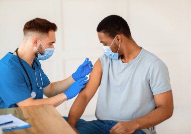 Tıbbi doktor ya da hemşire klinikteki Afrikalı Amerikalı erkek hastaya koronavirüs aşısı yapıyor. Yüz maskeli genç siyah adam covid-19 aşısı oluyor, aşı kampanyasına katılıyor.