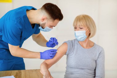 Maskeli yaşlı bir kadın koronavirüse karşı aşı oluyor, tıbbi yardım alıyor, klinikte bağışıklık kazanıyor. Erkek doktor, olgun kadın hastasına kovid 19 iğnesi yapıyor.