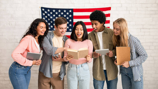 New study, learning and sharing knowledge. Surprised young multiracial students with notebooks and gadgets look in book in hands of asian woman, on white brick wall background with big USA flag