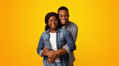 Mutlu Afro-Amerikan sevgililerinin portresi, sarılıp gülümseyen, sarı arka planda izole bir şekilde dikilen romantik siyahi adam, kız arkadaşına arkadan sarılıyor, boşluğu kopyalıyor.