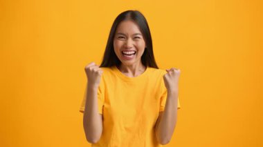 Bin yıllık Asyalı bir kadın mutluluğunu göstermek için gülümseyerek ve yumruklarını kaldırarak sevinçlerini ifade ediyor. İzole edilmiş, parlak sarı bir arka plana karşı duruyor, hayat dolu bir yaşam tarzını somutlaştırıyor..