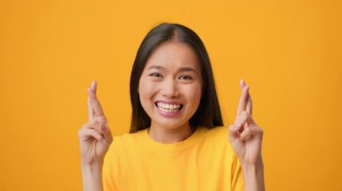 Milenyum Asyalı uzun saçlı bir kadın neşeli bir jest yapmak için parmaklarını çaprazlarken geniş bir gülümseme sergiliyor. Parlak sarı arka plan onun canlı ifadesini güçlendiriyor, pozitif enerji yayıyor..