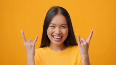 Bin yıllık Asyalı bir kadın neşeli bir el hareketi yaparken geniş bir gülümseme sergiliyor. Parlak sarı arka plan neşeli ve hayat dolu atmosferi arttırır, yaşam tarzı içeriği için mükemmeldir..