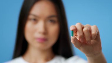 Bin yıllık Asyalı bir kadın uzun siyah saçlı elinde küçük yeşil bir kapsül taşıyor. Canlı, mavi bir arka plana karşı duruyor, merak ve nişan ifadesini vurguluyor..