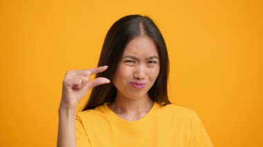 Bin yıllık uzun saçlı bir kadın, küçük bedenini gösteren bir jest yapıyor. Canlı turuncu bir arkaplanın önünde duruyor ve o ana enerji katıyor..