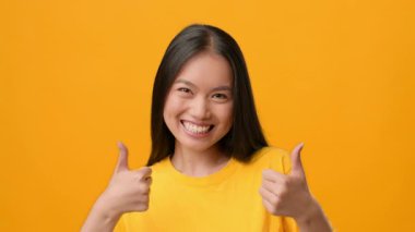 Genç bir Asyalı kadın parlak bir şekilde gülümserken parlak turuncu bir arka plana iki başparmak sallıyor. Neşeli ifadesi pozitiflik ve güven saçıyor, neşeli bir yaşam tarzını somutlaştırıyor..