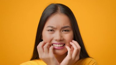 Uzun saçlı genç bir Asyalı kadın hayal kırıklığına uğramış bir yüz ifadesi sergiliyor, elleriyle yüzünü tutuyor. Parlak sarı arkaplan onun duygularını vurguluyor, yaşam tarzıyla ilgili bir anı yakalıyor..