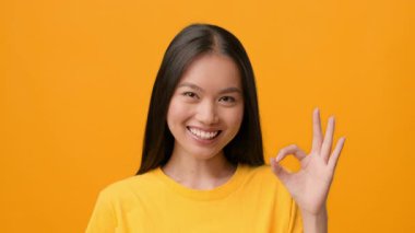 Sarı gömlekli neşeli bir Asyalı kadın sağ eliyle iyi bir jest yaparken gülümsüyor. Parlak sarı arka plan onun neşeli ifadesini ve gençlik enerjisini vurguluyor..