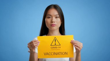 Genç bir Asyalı kadın elinde COVID-19 aşısının önemini vurgulayan parlak sarı bir tabela tutan mavi bir arkaplan karşısında duruyor. Ciddi ve odaklanmış görünüyor, farkındalığı arttırıyor..