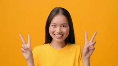 Uzun siyah saçlı neşeli bir kadın canlı sarı bir zemin üzerinde poz veriyor, iki eliyle de barış işareti gösteriyor. Onun parlak sarı gömleği canlı atmosferi tamamlar..