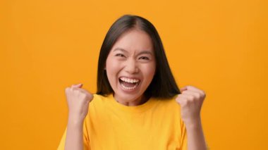 Uzun siyah saçlı, neşeli bir Asyalı kadın geniş bir şekilde gülümsüyor, kutlama için yumruklarını kaldırıyor. Canlı sarı arkaplan onun neşeli yüz ifadesini güçlendirir, canlı bir ruhu somutlaştırır..